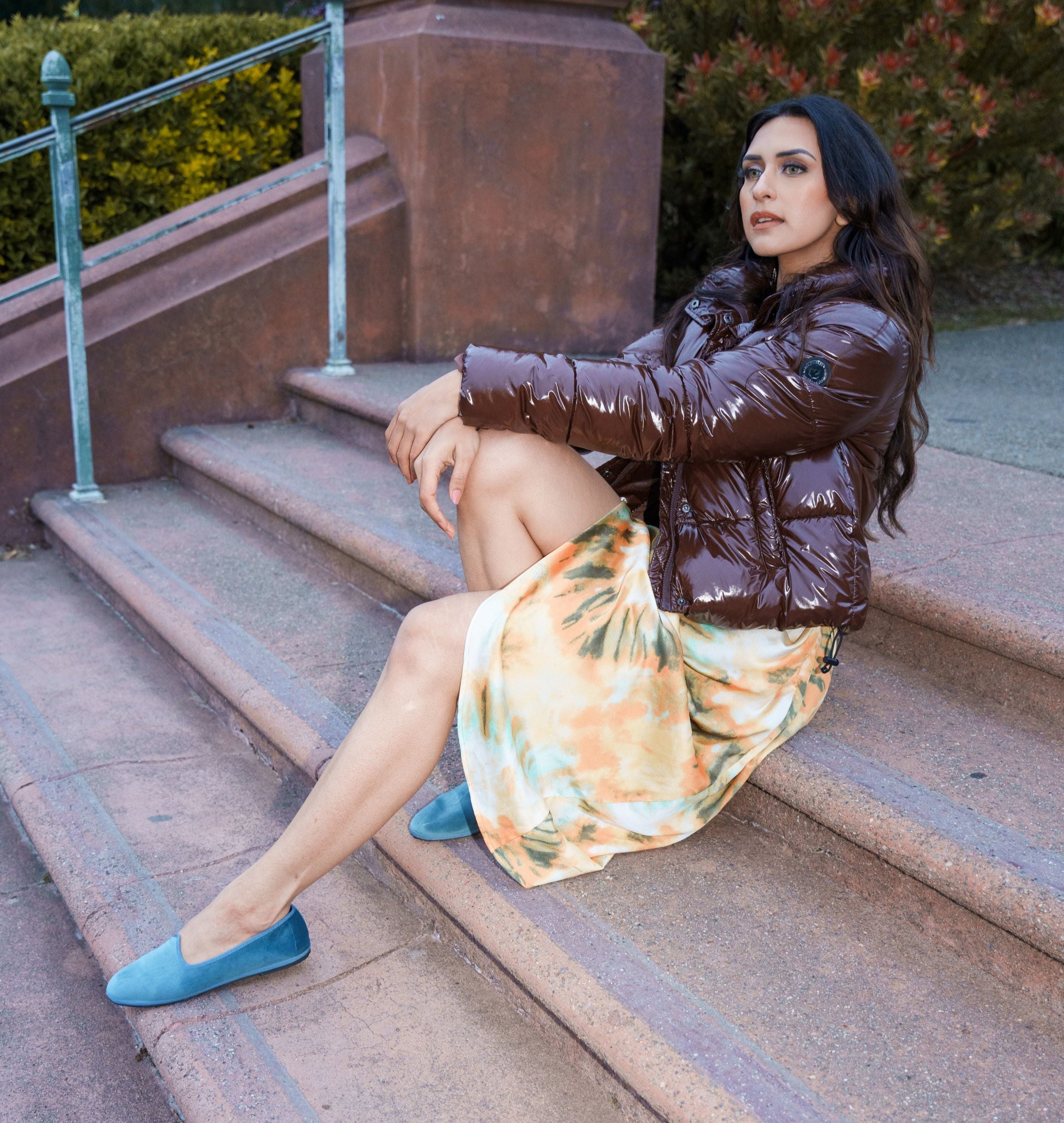 Woman sitting on steps wearing a brown jacket, colorful dress, and blue shoes. Fall style.