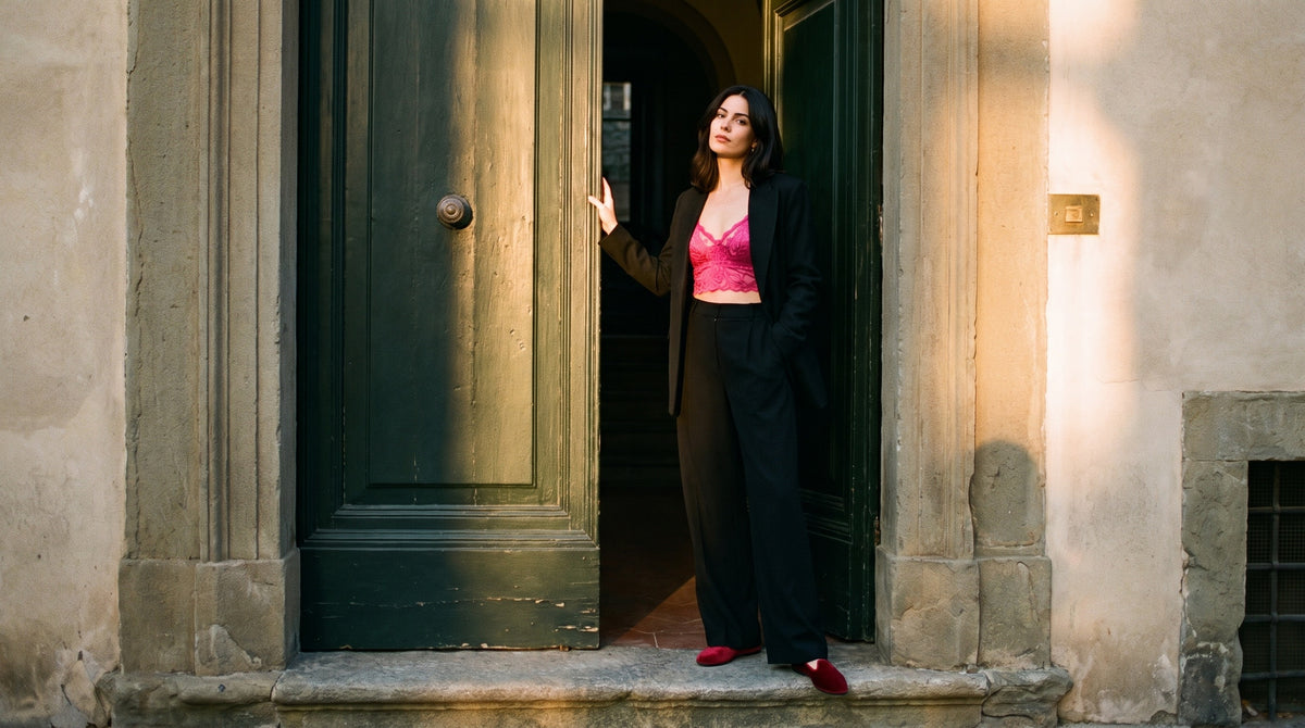 A woman stands in a doorway wearing a black blazer and wide-leg black trousers over a bright pink bralette, with red shoes, against a sunlit stone building.